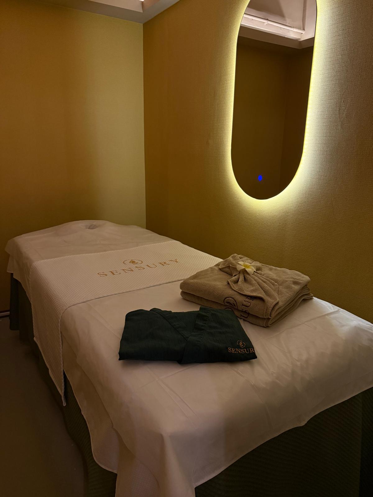 A Sensury treatment room with a bed, gold walls and a backlit oval mirror